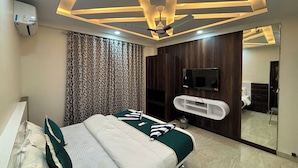 Habitación de hotel moderna con cama, televisor montado en la pared, ventilador de techo y baño visible a través de una puerta de cristal.