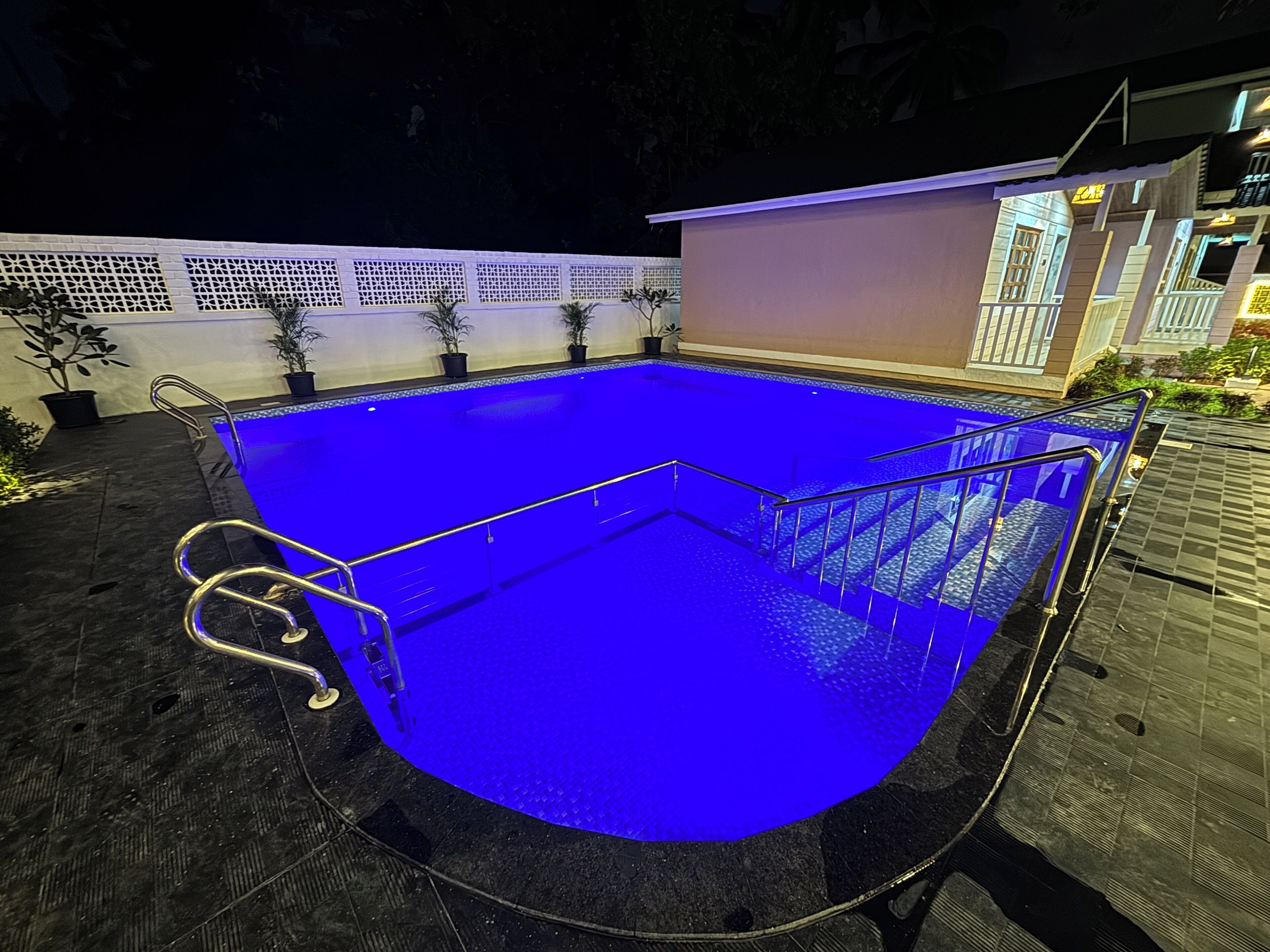 A swimming pool with a blue-lit interior, surrounded by a white fence and potted plants.
