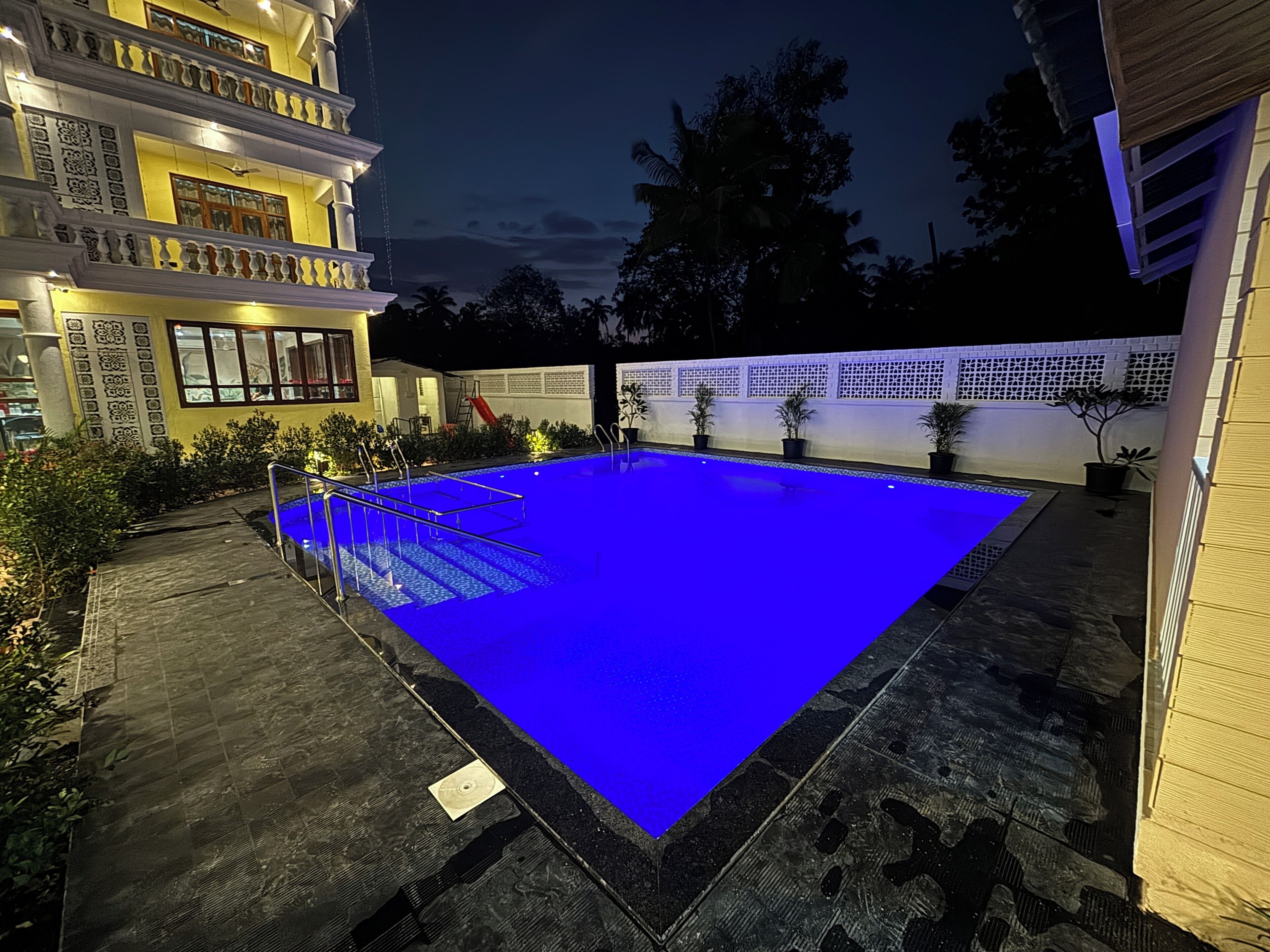 Une piscine dont le bord est illuminé de lumière bleue, entourée d’une terrasse dallée et de plantes en pot.