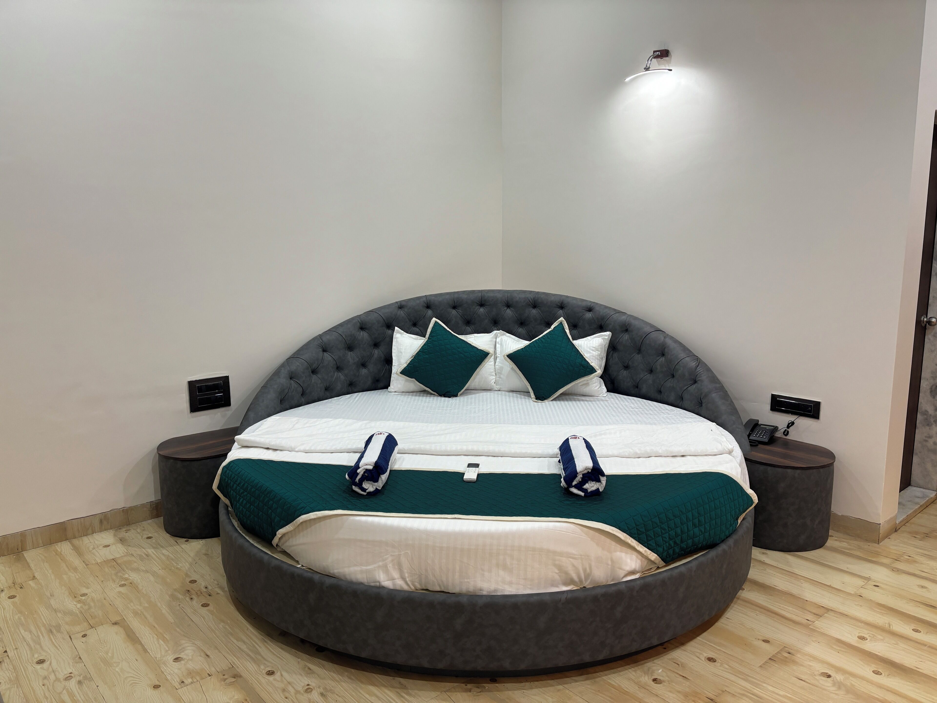 Ein modernes Schlafzimmer mit einem runden Bett, einem grau gepolsterten Kopfteil, weißer Bettwäsche und türkisfarbenen Kissen. Auf einem Nachttisch stehen eine Lampe und ein Telefon.