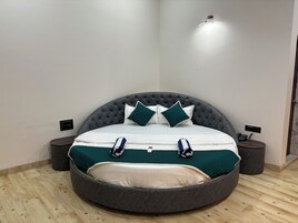 Un dormitorio moderno con cama redonda, cabecero tapizado en gris, ropa de cama blanca y almohadas color turquesa. Hay dos mesitas de noche, una con lámparas y un teléfono.