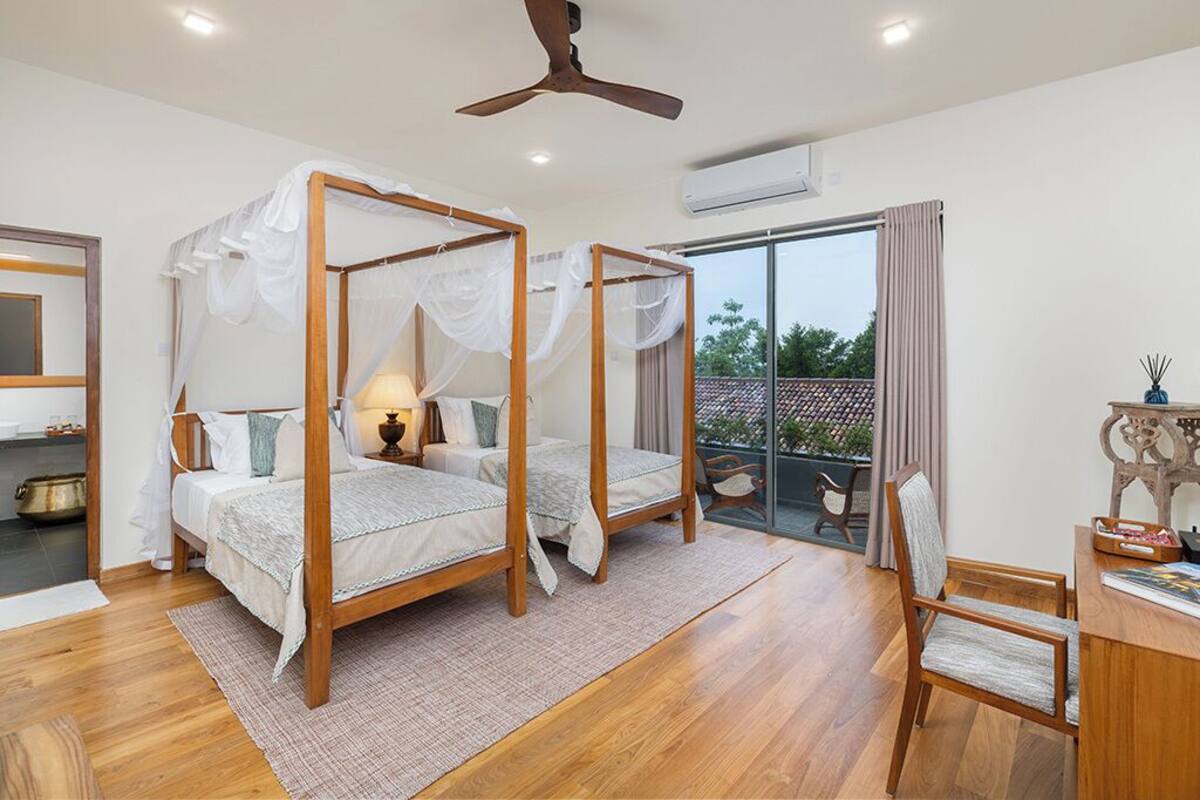 A bedroom with two four-poster beds, a desk with a chair, and a balcony with a view.