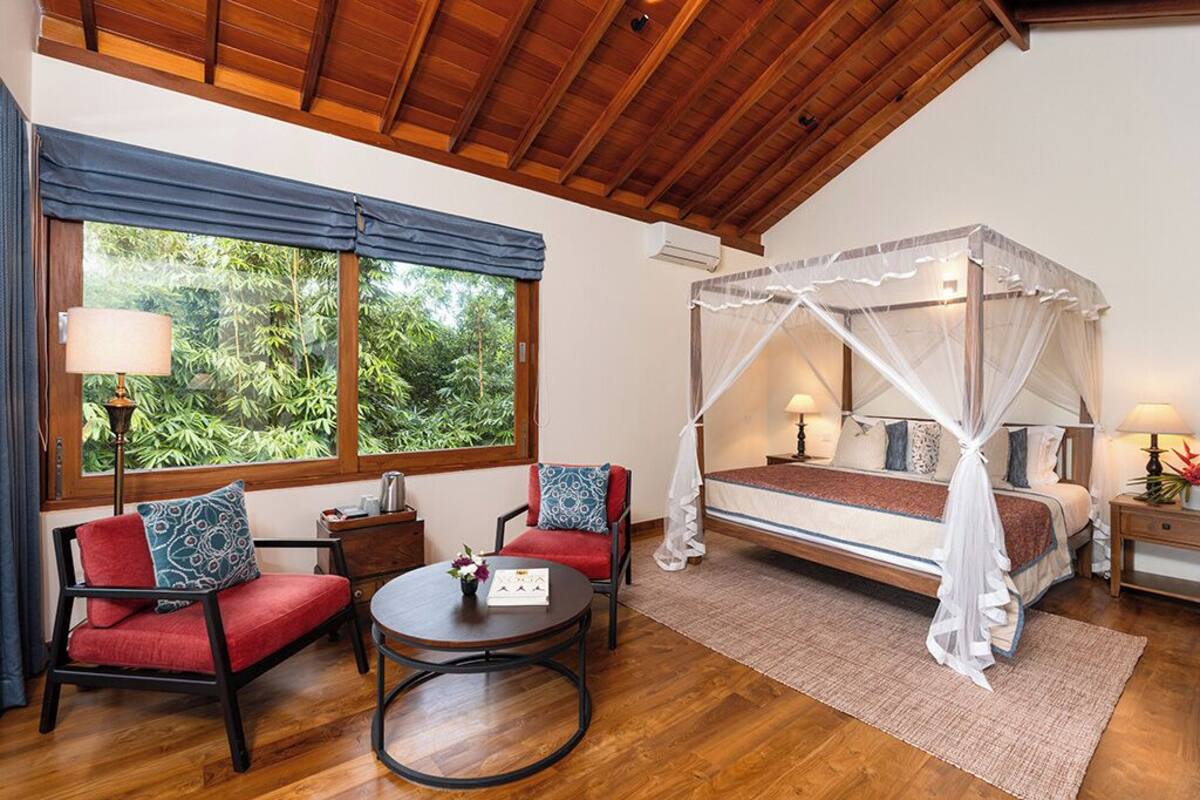 A bedroom with a canopy bed, a sitting area with a table and chairs, and large windows with curtains.
