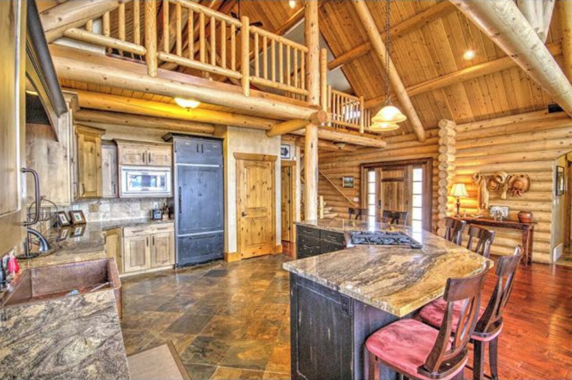 A log cabin interior with a kitchen, dining area, and a loft with a balcony.