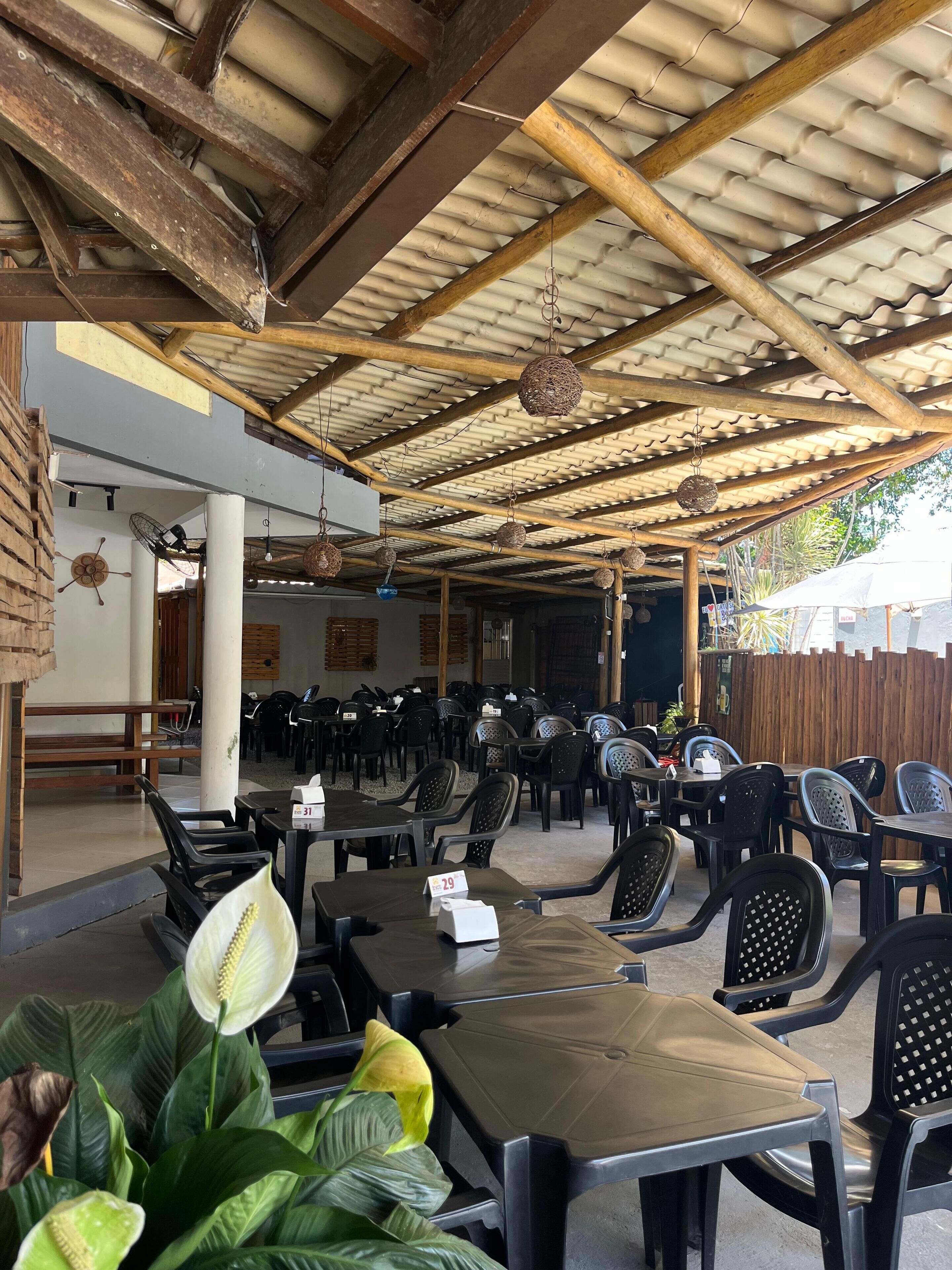 Un espace extérieur abrité, aménagé avec des tables et des chaises, doté d’un plafond en bois et de luminaires suspendus.