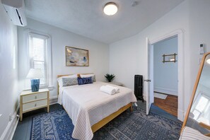 Ein Schlafzimmer mit einem Bett, einem Nachttisch, einer Lampe, einem Gemälde und einer Tür zu einem anderen Raum.