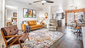 Un salon spacieux doté d’un grand ventilateur de plafond, d’un canapé en cuir marron, d’un tapis à motifs et d’un mur décoratif en briques.