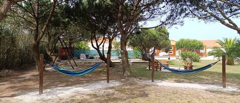 A hammock between trees in a park area with a playground and buildings in the background.