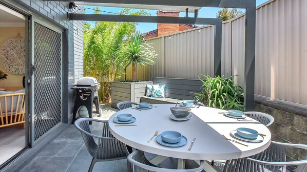 Enjoy dining al fresco with a round table set for four, surrounded by a grill, lush greenery, and a residential view.