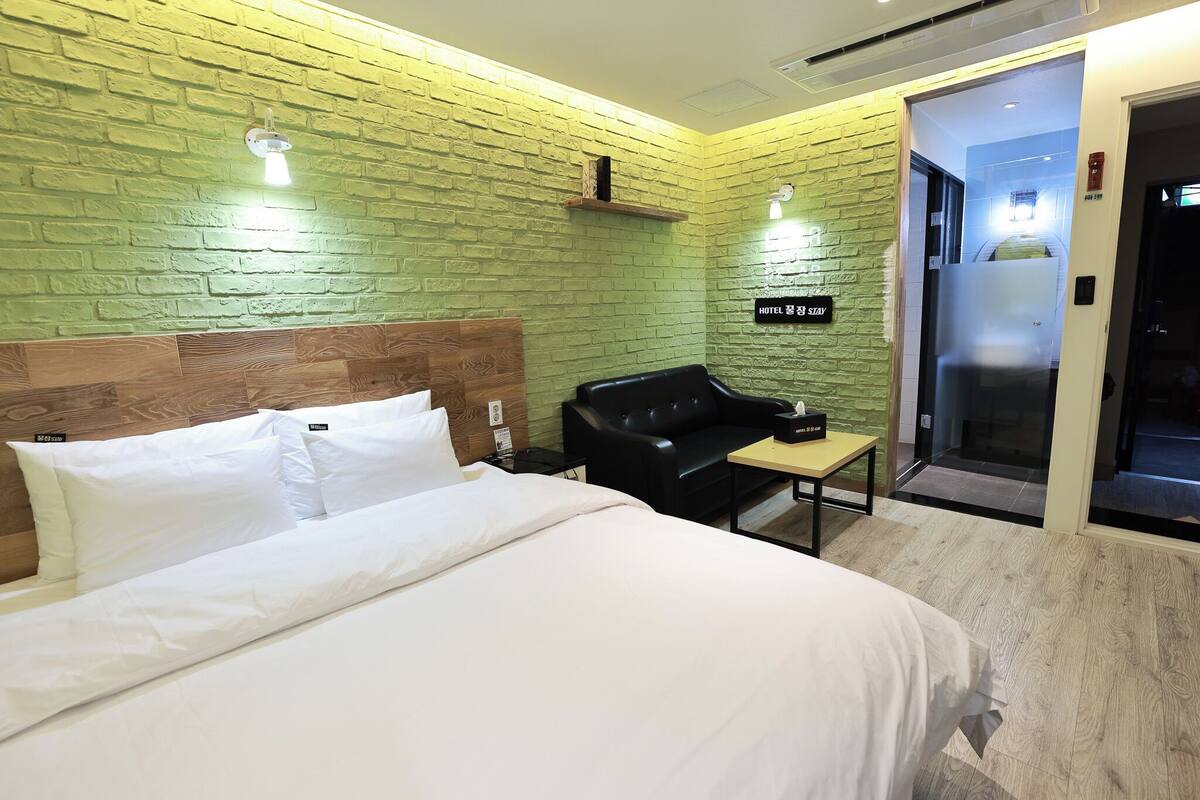 Een hotelkamer met een groene bakstenen muur, een houten hoofdbord, een zwarte leren bank, een klein tafeltje en een bed met witte lakens.