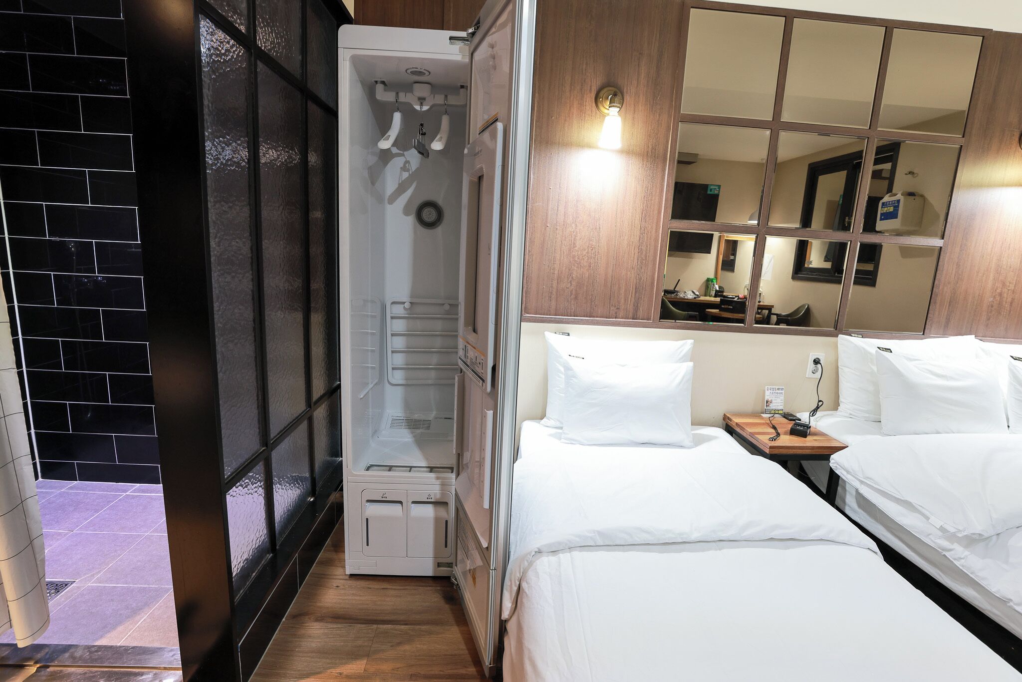 Una habitación de hotel con vestidor, una cama con ropa de cama blanca, una mesita de noche de madera y un espejo grande que refleja la habitación.