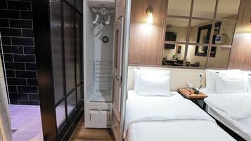 Una habitación de hotel con vestidor, una cama con ropa de cama blanca, una mesita de noche de madera y un espejo grande que refleja la habitación.