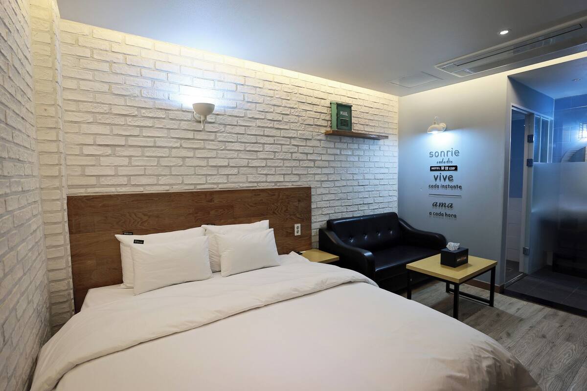 Een hotelkamer met een witte bakstenen muur, een houten hoofdeinde, een bed met witte lakens, een zwarte leren bank en een klein tafeltje met een zakdoos.