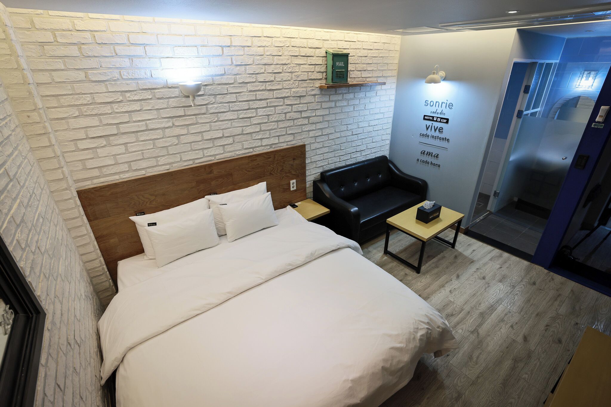 A hotel room with a brick-patterned wall, a wooden headboard, a bed with white linens, a black leather sofa, a small table, and a wall-mounted shelf with a mail slot.