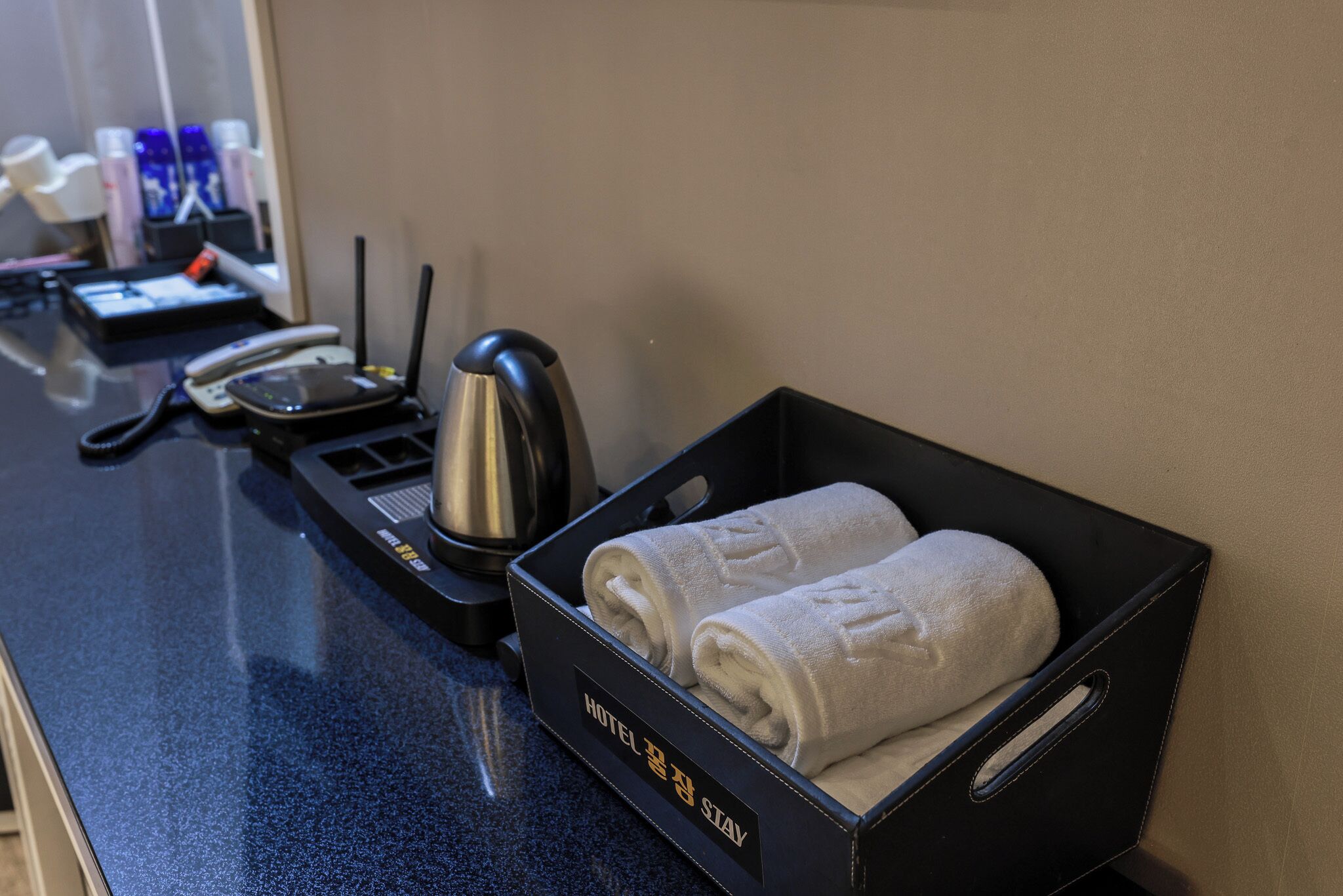 Mostrador de hotel con una bandeja negra que contiene dos toallas enrolladas, una tetera, un teléfono y una cafetera.