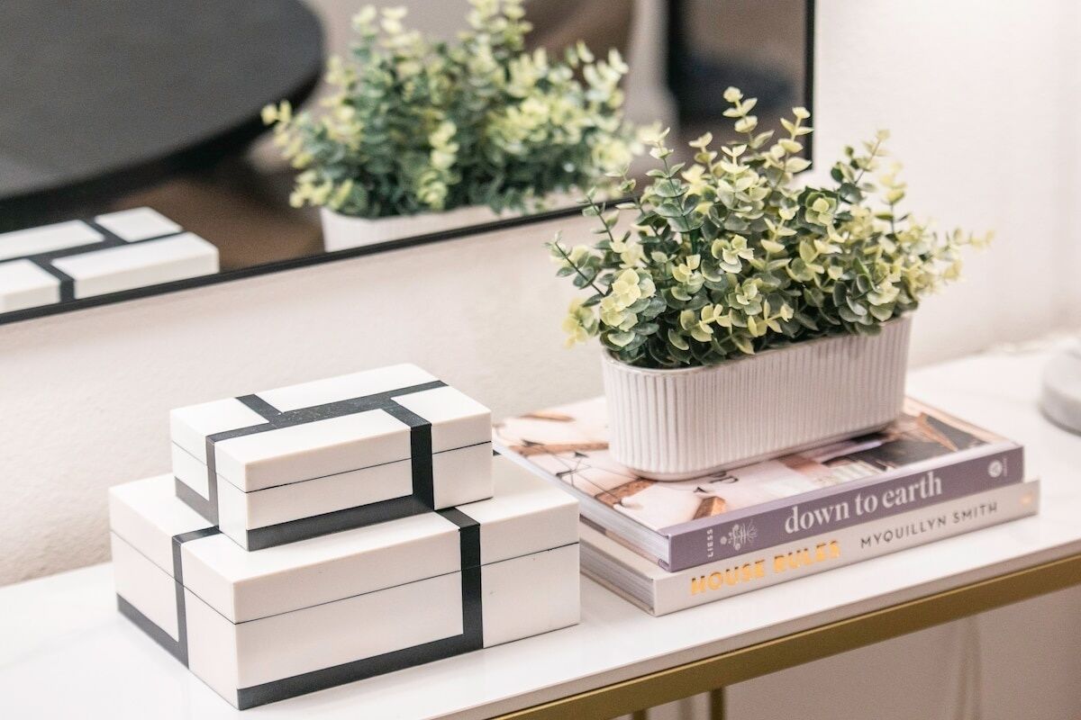 Un escritorio blanco con una pequeña planta en maceta, una pila de cajas blancas y un libro titulado 'Down to Earth' de Myquillyn Smith.