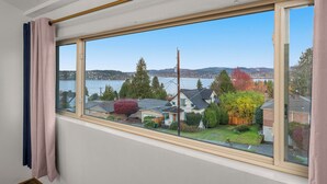Ein Zimmer mit großem Fenster und Blick auf eine Wohngegend mit Häusern, Bäumen und einem Gewässer.