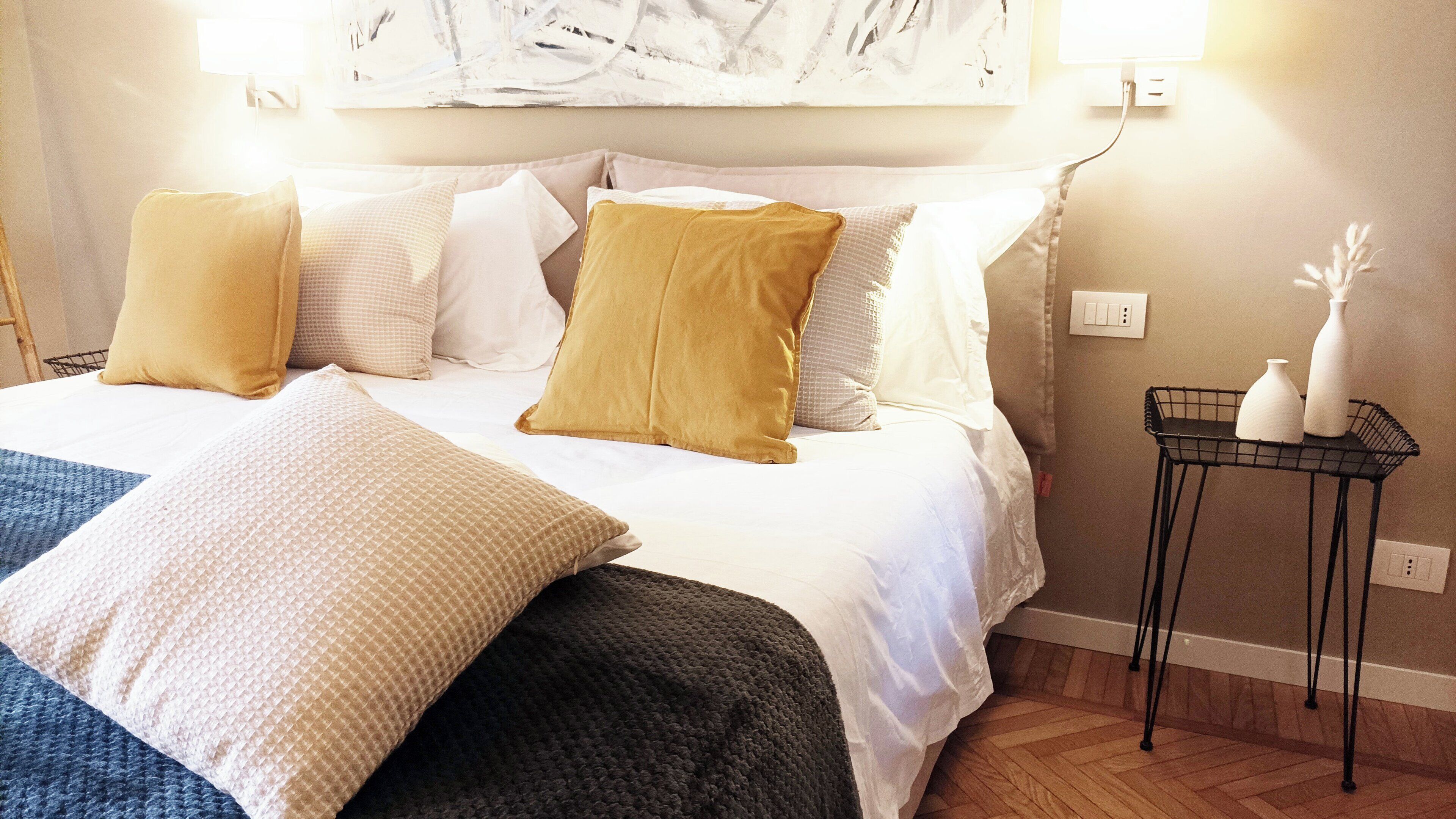 A neatly made bed with multiple pillows and a black metal side table with two white vases.