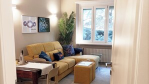 A living room with a sofa, ottoman, and a dining area with a table and chairs. There is a large plant and artwork on the wall. - QuickHome - Le due Palme (Biella)