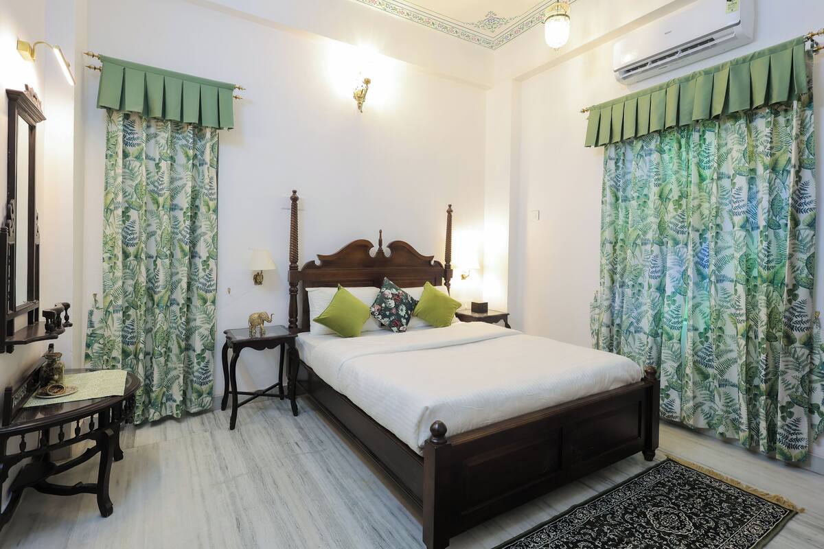 A bedroom with a wooden bed, green curtains, a bedside table, a lamp, and an air conditioning unit.