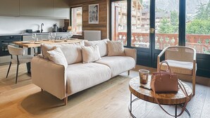 Een moderne woonkamer met een bank, salontafel en stoelen. Er is een keuken met een eettafel en stoelen. Grote ramen bieden uitzicht op een balkon met tuinmeubilair.