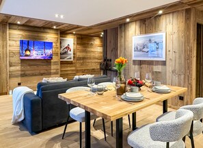 Una sala de estar moderna con interior de madera, un sofá, una mesa de comedor preparada para cuatro personas y un televisor montado en la pared.