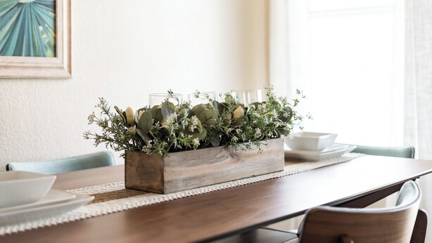 Una mesa de comedor de madera con un centro de decoración de follaje y velas, sobre un mantel individual blanco.