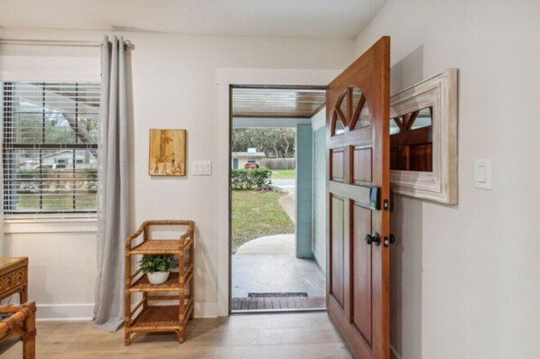 Une porte-fenêtre donnant sur un espace extérieur abrité, avec une porte en bois, une fenêtre équipée de stores et une étagère en osier sur laquelle est posée une plante.
