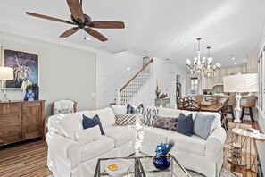 A spacious living room with a white sofa set, wooden flooring, a ceiling fan, and a dining area with a chandelier. - Seagrove Gardens Mins to the Beach, Santa Rosa Retreat w/ A+ Design (Santa Rosa Beach)