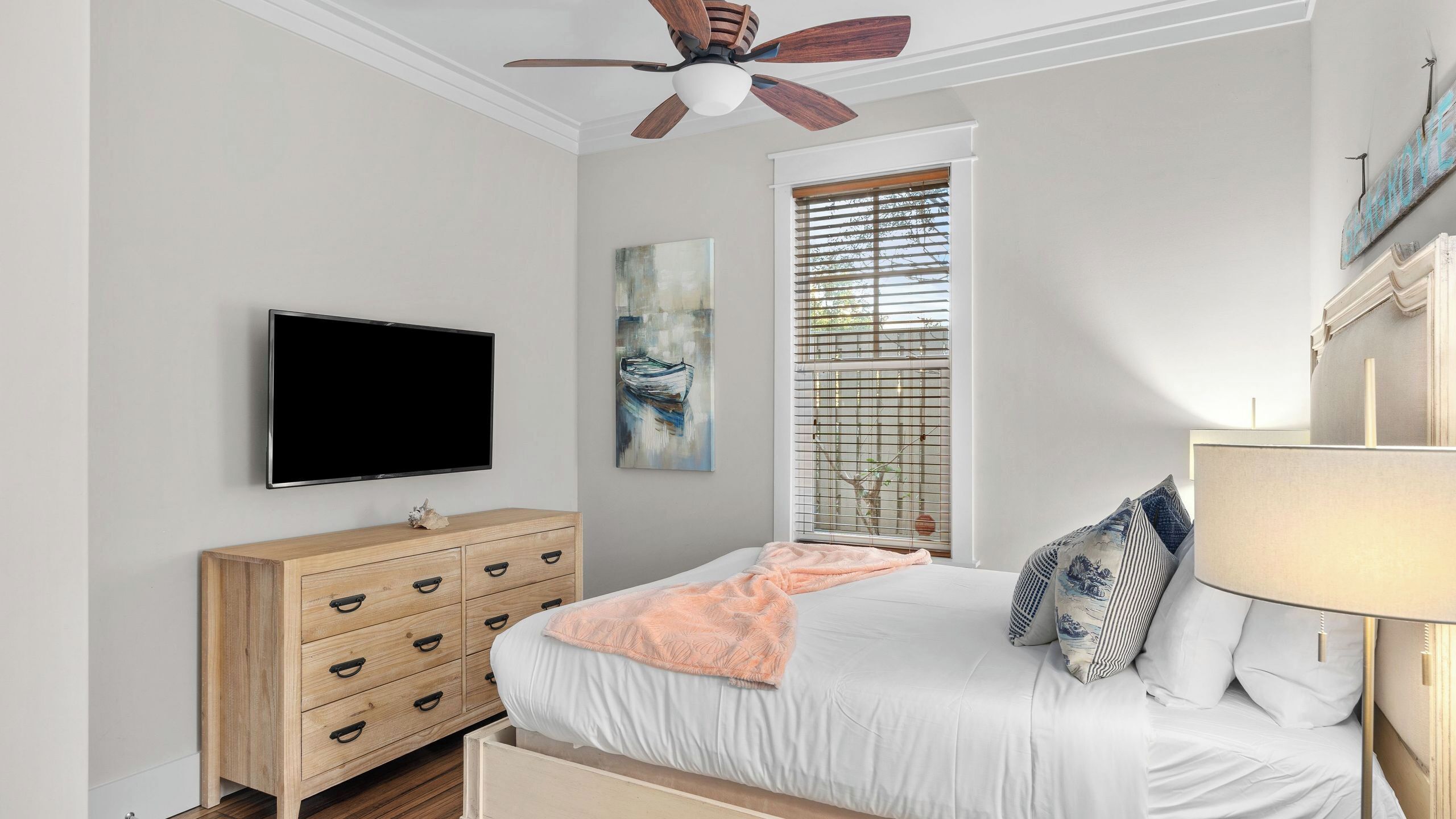 A bedroom with a bed, a TV, a wooden dresser, a ceiling fan, a window with blinds, and a painting.