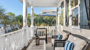 Un porche cubierto con barandillas blancas, muebles de mimbre y vistas a una casa y palmeras.