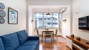 A modern living room with a blue sofa, a wooden dining table with chairs, a TV mounted on the wall, and a large window with a city view. - Refuge in Copacabana 5min From the Beach Nsc1102 (Rio de Janeiro)