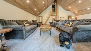 A spacious loft with high ceilings, exposed wooden beams, and a central staircase. There are two grey sofas with decorative pillows and a wooden coffee table in the center. A black teddy bear is on the floor near the sofas. - 20+ Guests-20 Minutes to Yellowstone-Hot Tub-Family Reunions-Fly Fishing (Island Park)