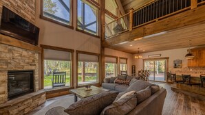 A spacious living room with a stone fireplace, large windows, a high ceiling, and a balcony. - 20+ Guests-20 Minutes to Yellowstone-Hot Tub-Family Reunions-Fly Fishing (Island Park)