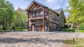 A large log cabin with a balcony and a garage, surrounded by a forested area. - 20+ Guests-20 Minutes to Yellowstone-Hot Tub-Family Reunions-Fly Fishing (Island Park)