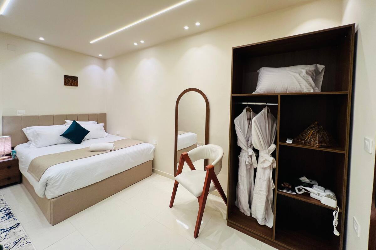 Ein modernes Hotelzimmer mit einem ordentlich bezogenen Bett, einem Holzstuhl, einem Bademantelständer mit Handtüchern und einem Nachttisch mit einer Lampe.