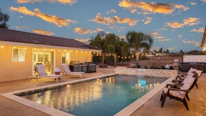 Un patio trasero con piscina, muebles de exterior, una parrilla y luces colgantes.