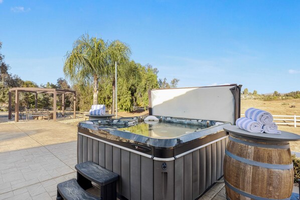 Un bain à remous en forme de tonneau en bois, avec des serviettes à proximité, entouré de meubles d’extérieur et d’une pergola.