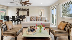 A living room with a sofa, two armchairs, a coffee table with drinks and flowers, a dining area with chairs, and a ceiling fan. - Bordeaux by Avantstay Private Pool, Hot Tub & Game Room Near Temecula (Temecula)