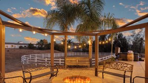 A covered outdoor seating area with a fire pit, benches, string lights, and a view of a fenced field. - Bordeaux by Avantstay Private Pool, Hot Tub & Game Room Near Temecula (Temecula)