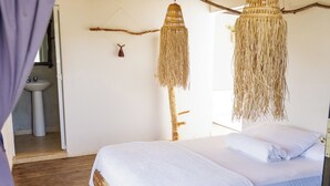 Une chambre avec un lit en bois, du linge de lit blanc, un luminaire suspendu en fibres tressées, et une salle de bain visible, équipée d’un lavabo et d’un miroir.