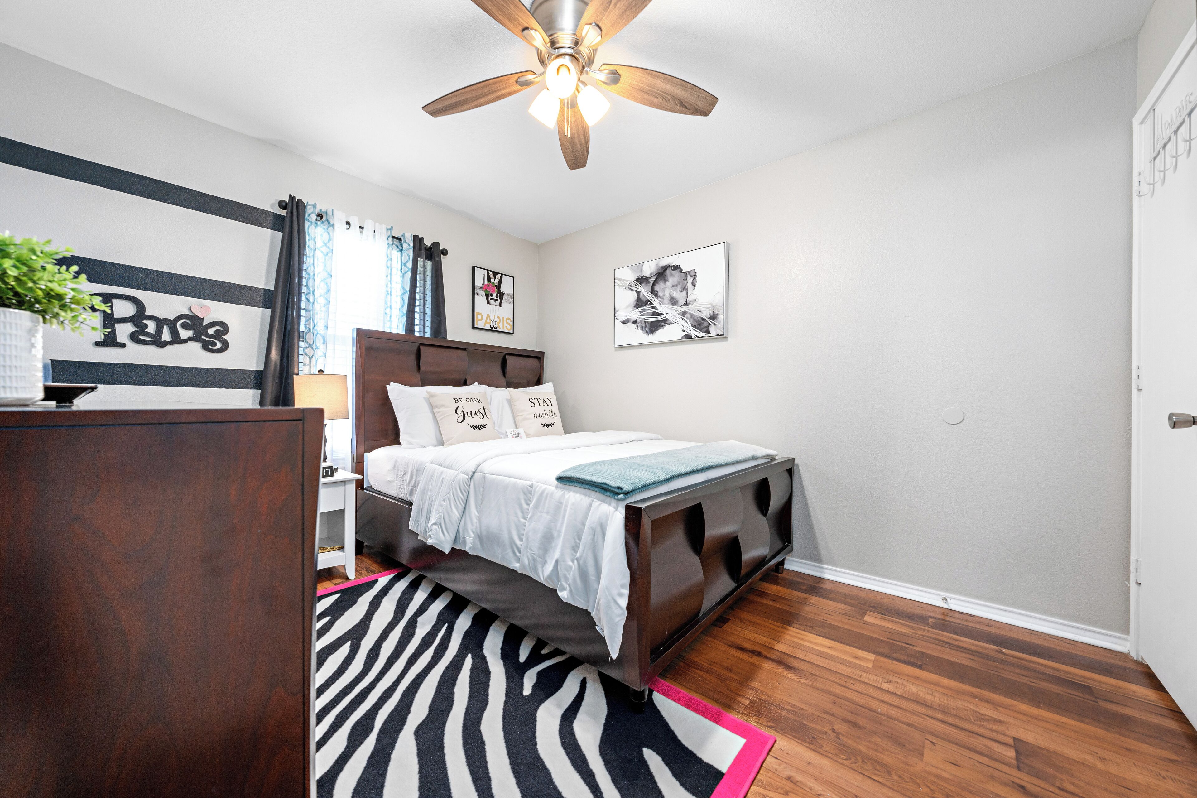 A bedroom with a bed, a ceiling fan, a zebra-patterned rug, and a wall with a 'Paris' sign.