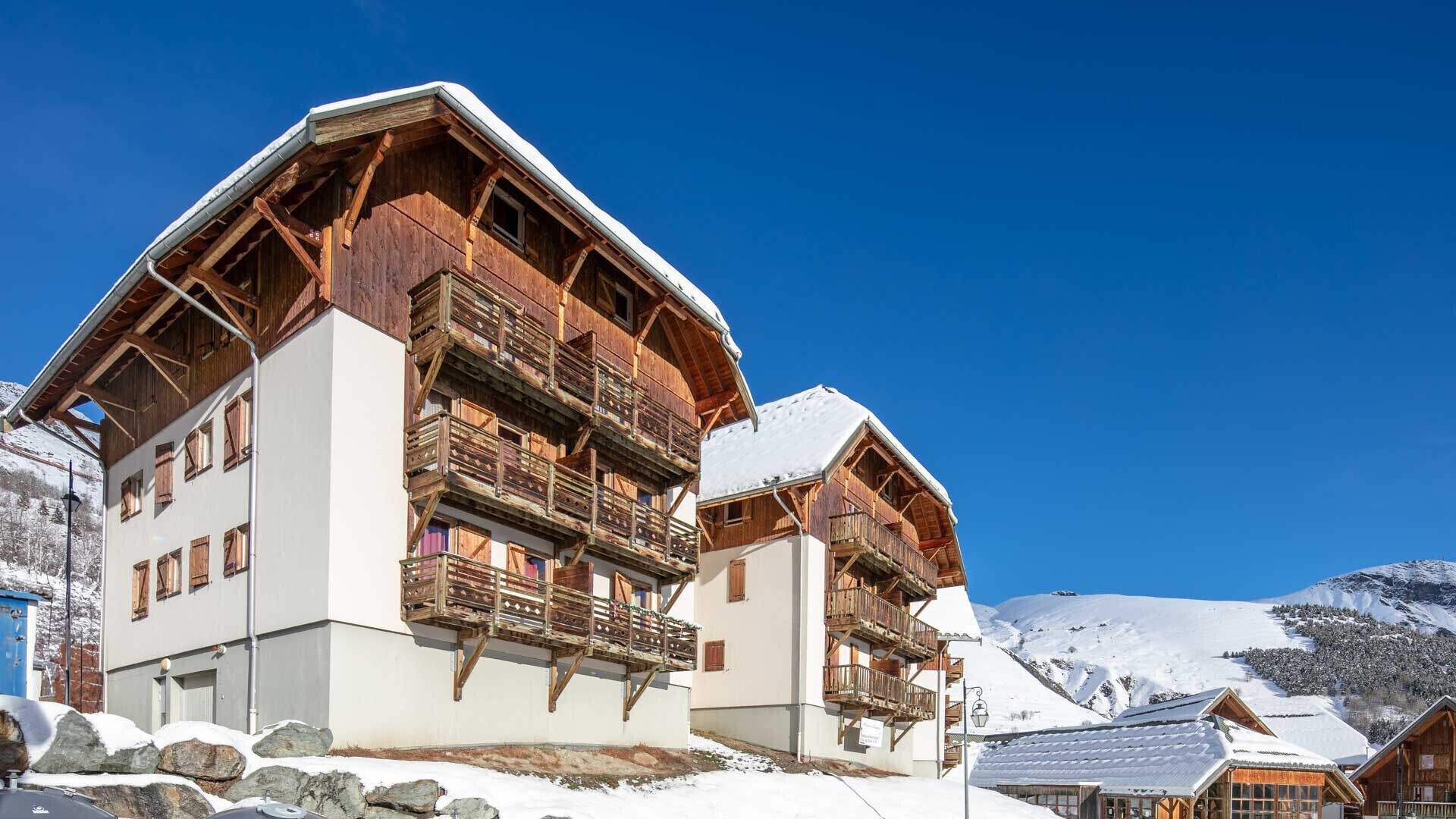 Ein Chalet mit Holzbalkonen und einem mit Schnee bedeckten Dach, vor einer Bergkulisse.