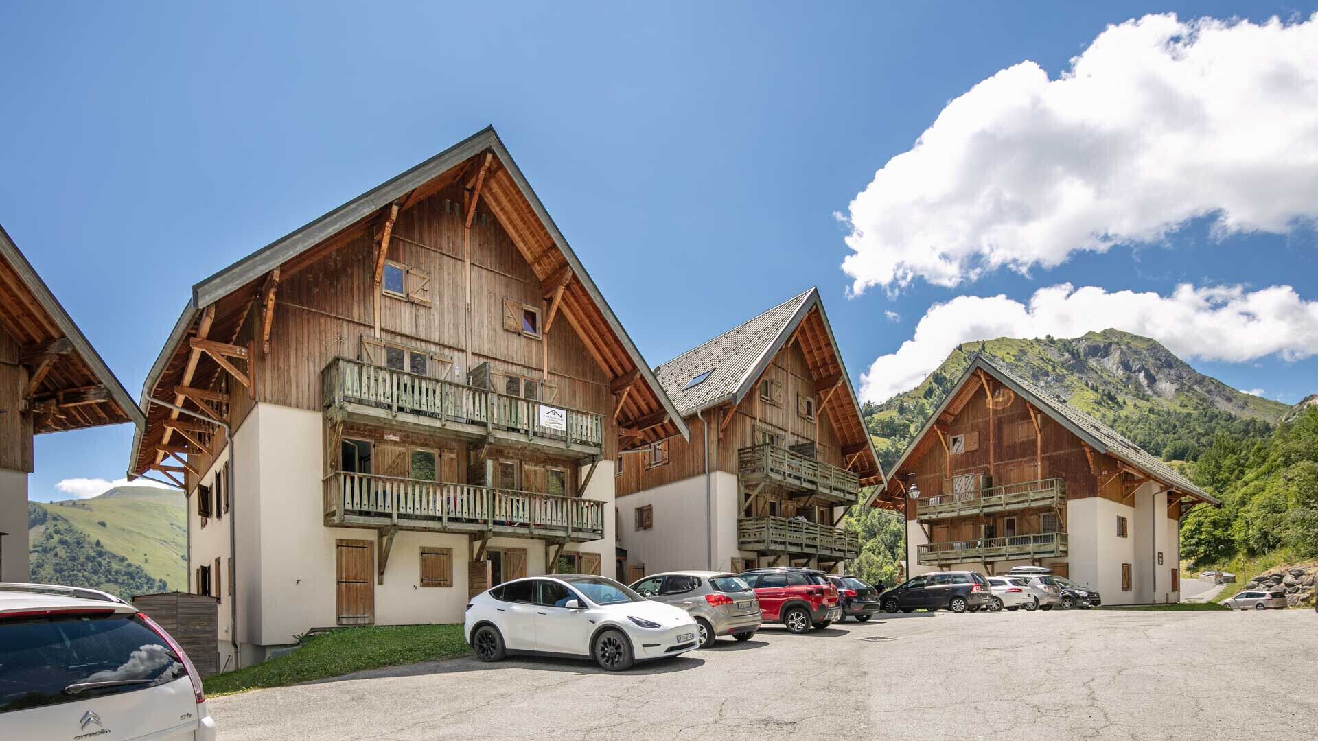 A row of wooden chalet-style buildings with balconies, a parking lot, and a mountainous backdrop.