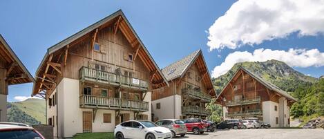 A row of wooden chalet-style buildings with balconies, a parking lot, and a mountainous backdrop.