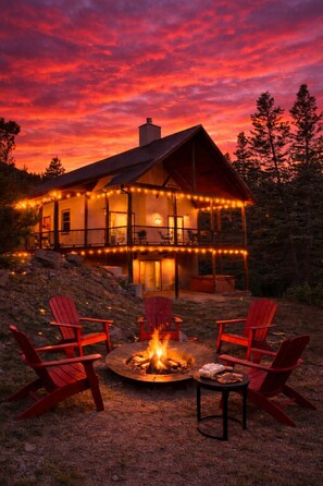 A cabin with a fire pit and red chairs, surrounded by a deck with string lights.