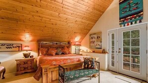A cozy bedroom with a wooden ceiling, a large bed with a red bedspread, a wooden nightstand, a bench, and a door leading to a balcony.