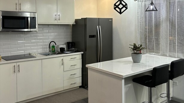 Een moderne keuken met witte kasten, een zwarte koelkast, een magnetron en een eethoek met twee zwarte barkrukken.