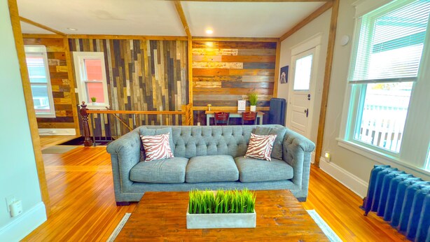 Una sala de estar con un sofá gris, suelo de madera, una mesa de centro con una planta, una pared de madera con un banco y una ventana con persianas.
