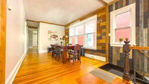 Un comedor con una mesa y sillas de madera, ventanas amplias con persianas y una escalera con barandilla de madera.